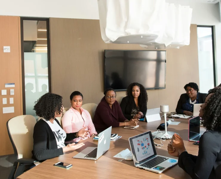 A group of people, including several women, sitting around a conference table with laptops, having a meeting in a modern office with large windows and a TV on the wall.