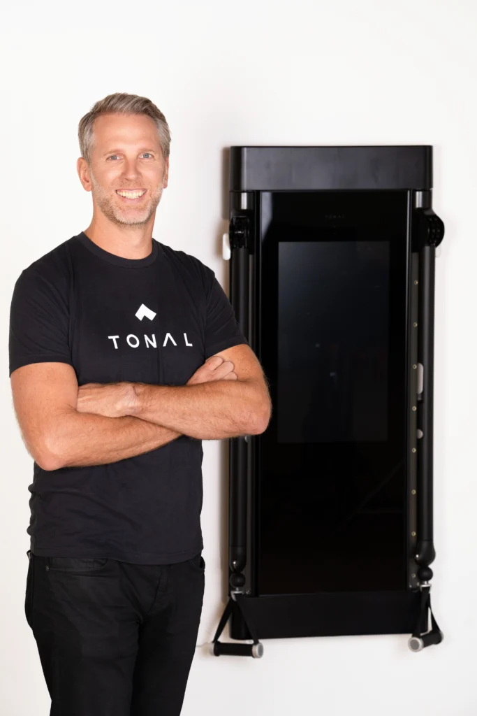 a smiling image of new Tonal CEO Darren MacDonald next to a Tonal smart home gym.