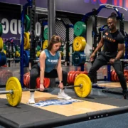 woman picks a barbell up off the ground next to her personal trainer