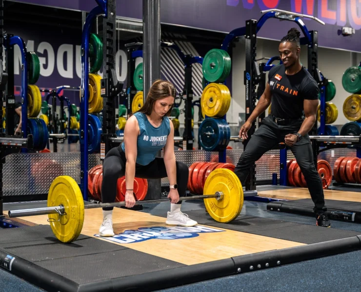 woman picks a barbell up off the ground next to her personal trainer