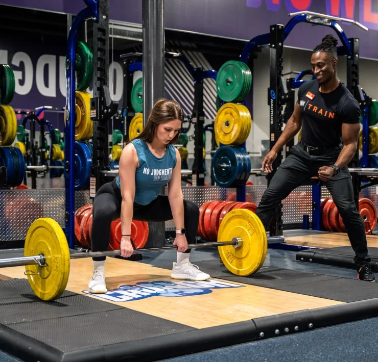 woman picks a barbell up off the ground next to her personal trainer